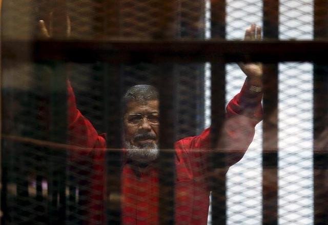 Deposed President Mohamed Mursi greets his lawyers and people from behind bars at a court wearing the red uniform of a prisoner sentenced to death, during his court appearance with Muslim Brotherhood members on the outskirts of Cairo, Egypt, June 21, 2015. REUTERS/Amr Abdallah Dalsh/File Photo